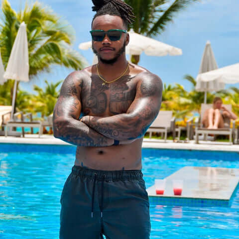 FTM transgender person with tattoos and sunglasses standing shirtless by a pool in a tropical resort, wearing dark swim shorts, with arms crossed. Palm trees, umbrellas, and lounge chairs in the background.