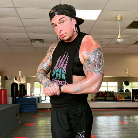 FTM transgender person with tattoos posing in a gym, wearing a black sleeveless shirt and cap turned backward. They have stretched earlobe piercings and are standing with arms slightly flexed, with mirrors, mats, and gym equipment in the background.