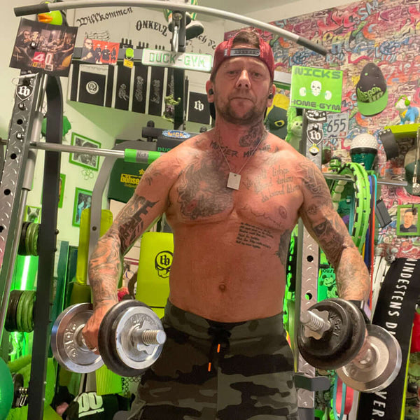 FTM transgender person lifting dumbbells shirtless in a colorful home gym. They have multiple tattoos on chest and arms, wear a red backward cap and camouflage shorts, surrounded by green gym equipment and wall decorations.