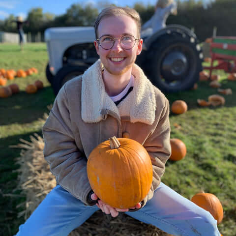FTM transgender person smiling and holding a pumpkin while sitting on a hay bale in a pumpkin patch. Wearing glasses and a tan shearling jacket, with scattered pumpkins and a tractor in the background.